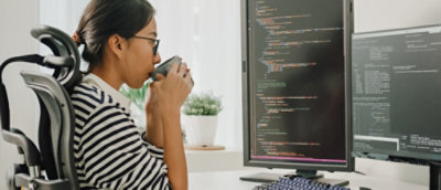 A person in a striped shirt is sitting at a desk, viewing code on a vertical monitor while sipping from a mug.