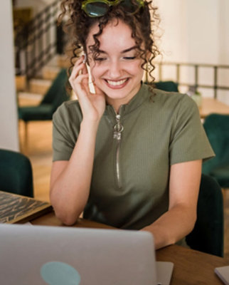 A smiling woman with curly hair, in a green top, sits at a table using a laptop and talking on the phone.