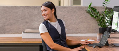 A women sitting at a desk with a monitor, plant, and papers in an office setting.
