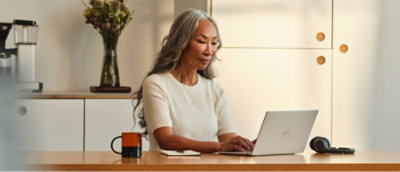 A women with long gray hair using a laptop at a table with a coffee mug and headphones.