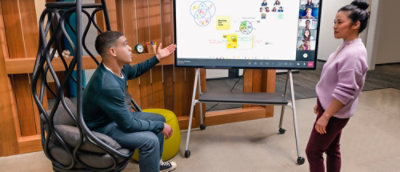 Two colleagues discussing a presentation displayed on a large screen during hybrid meeting.