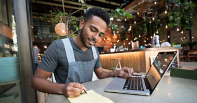 Muškarac sedi za stolom na kojem se nalazi laptop i piše u beležnicu u dobro osvetljenom kafiću sa visećim biljkama i ukrasnim izloženim sijalicama.