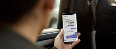A person in a car looking at a smartphone displaying a messaging app with multiple conversations visible.