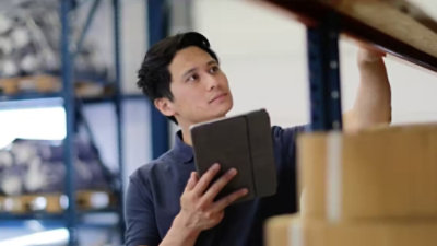 Person in a warehouse holding a tablet and reaching for a box on a shelf, with storage items visible in the background.