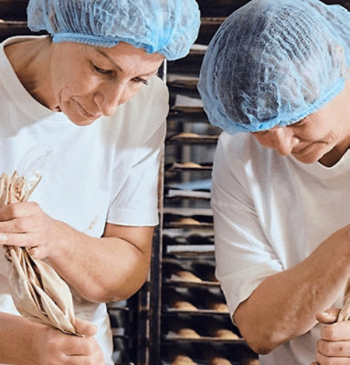 Two individuals in white uniforms and blue hairnets holding baked goods in front of racks filled with similar items.