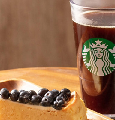 Slice of cheesecake topped with blueberries on a wooden plate beside a glass of Starbucks coffee