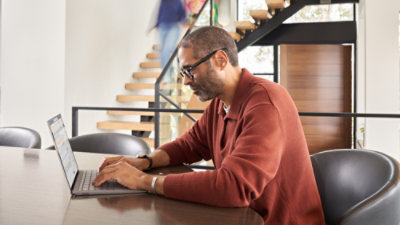 A person working on a laptop at a table in a modern indoor space with a staircase and railing in the background.