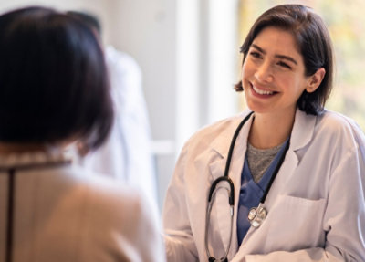 A doctor wearing a white lab coat and stethoscope speaks with another person in a clinical setting.
