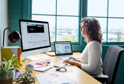 Person working at a desk with a desktop monitor and laptop near a window.