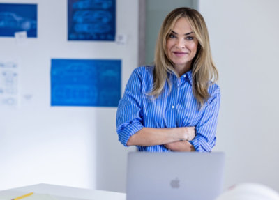 Person standing behind a laptop with arms crossed, blueprints on wall, modern workspace setting.