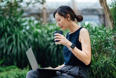 A person sitting on a bench using a laptop and holding a cup.