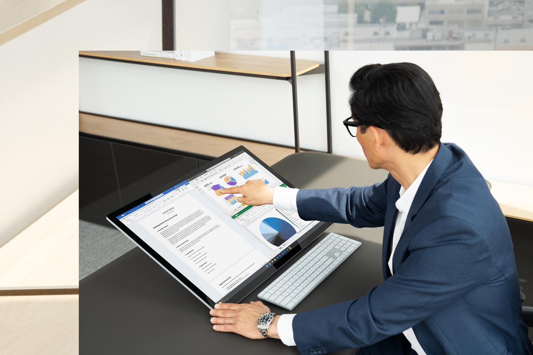 Sitting at his desk, a man touches the screen of Surface Studio 2+ making a business presentation with the screen titled back to studio mode.