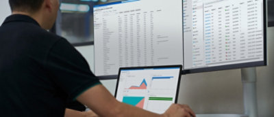 Osoba u tamnoj košulji posmatra podatke na više ekrana računara i laptopu u modernom kancelarijskom okruženju.