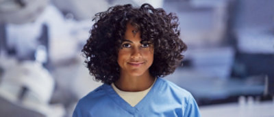 Healthcare professional in blue scrubs standing in a clinical environment with medical equipment in the background.