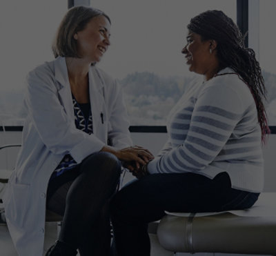 Two people seated together in a consultation setting, one offering support while speaking with the other near a window.