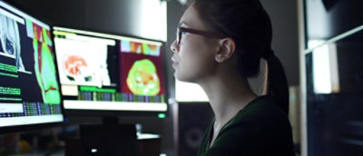 Medical researcher reviewing diagnostic images on multiple computer monitors in a dimly lit workspace.