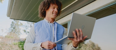 Person standing outdoors using a tablet and laptop near a building entrance.