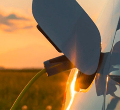 Close-up of an electric vehicle being charged at sunset, with the cable plugged into the port and a natural field in the background.