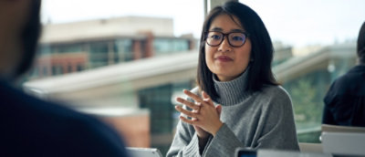 Uma mulher usando óculos e uma blusa com gola alta cinza com as mãos juntas.