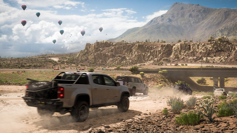 Forza Horizon 5. Trucks race down a dirt road in Mexico with hot air balloons in the distance. 