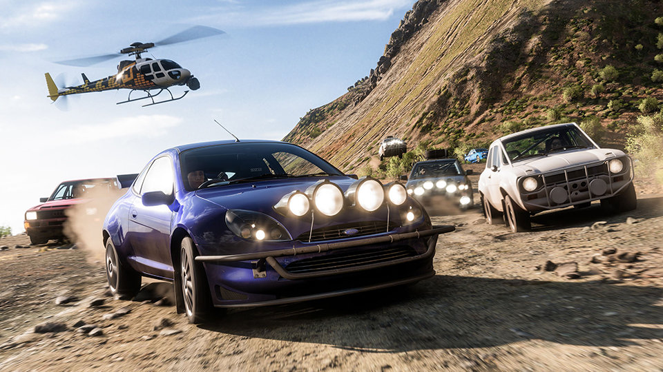 Forza Horizon 5. Une Ford Puma conduisant un groupe de voitures sur un chemin de terre avec un hélicoptère en vol stationnaire au loin.