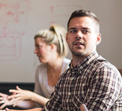 Two people in a discussion setting, with one gesturing and a whiteboard in the background.