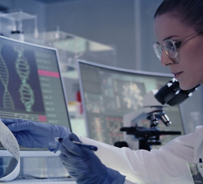 Person in a laboratory wearing protective eyewear and gloves, writing notes while viewing DNA sequences on computer screens with a microscope in the background.