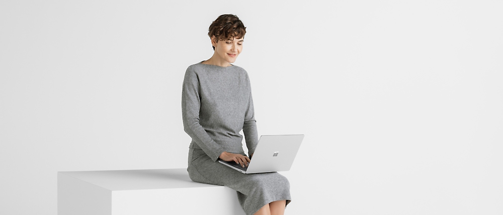 Kobieta w szarej sukience, która używa laptopa, siedząca na białej ławce na tle gładkiego białego tła.
