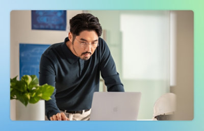A man wearing a black shirt looking at a laptop with a plant in a pot nearby.