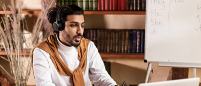 A man wearing headphones sitting near a whiteboard with writing on it and a bookshelf in the background.