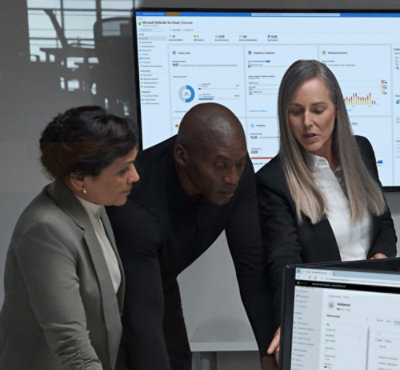 Three people reviewing data on laptops with an analytics dashboard displayed on a screen.
