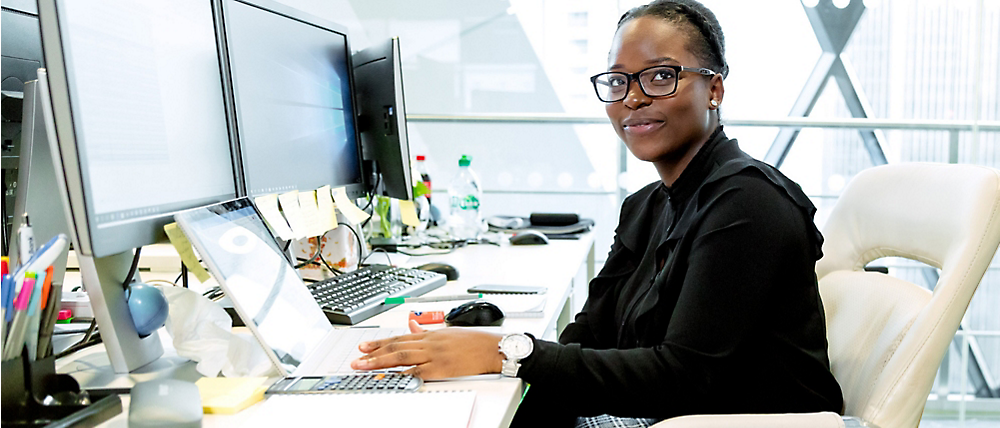 صورة لامرأة ترتدي نظارة وقميصاً أسود تجلس على مكتب به شاشات كمبيوتر متعددة في مكتب مضاء جيداً.