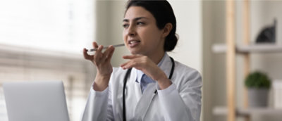 A doctor in a white coat uses a voice recording function on a smartphone while seated at a desk with a laptop.