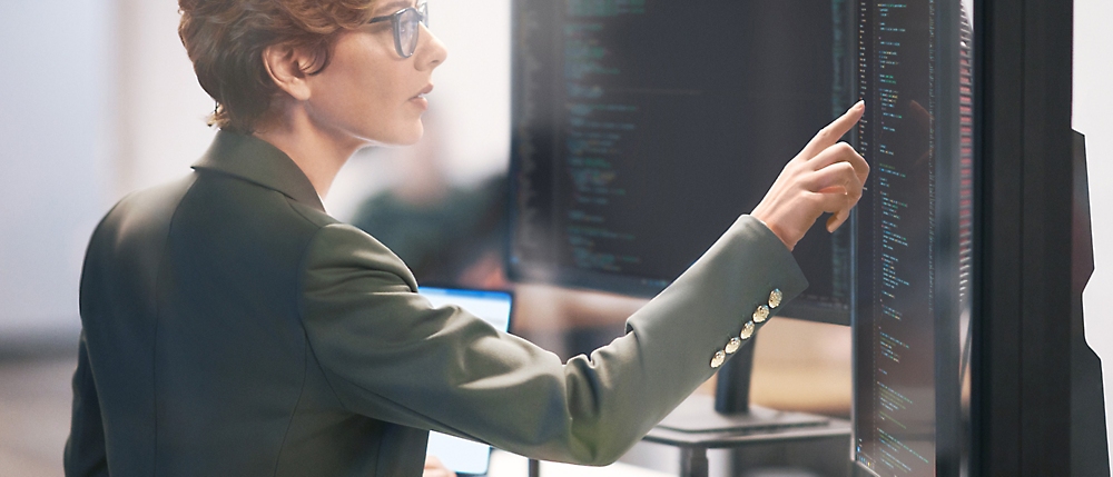 Una mujer profesional con una chaqueta oscura examina y señala datos en varias pantallas de equipos en un entorno de oficina.