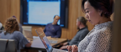 A team meeting where one person works on a laptop while others discuss beside a screen.