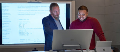 Two colleagues review code on a large monitor using a Surface device in a meeting room.