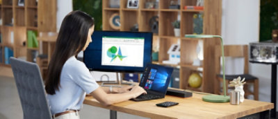 A woman works at a desk on a laptop connected to a monitor displaying graphs, in a modern office with shelving units in the background.