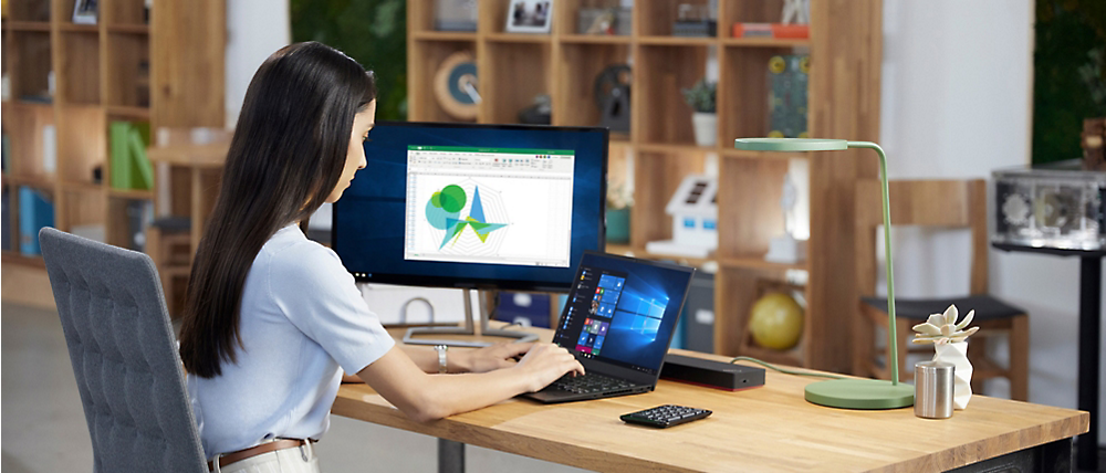 Жінка працює за столом на ноутбуці, підключеному до монітора з діаграмами, у сучасному офісі зі стелажами на задньому плані.