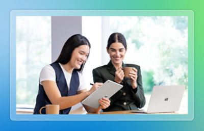 Two women smiling while working together on a tablet and laptop, one holding a coffee mug in a bright office setting.