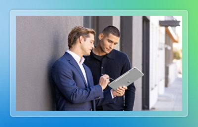 Two men standing outdoors by a wall, reviewing content on a tablet together, one using a stylus while the other looks on.