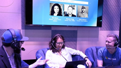 Three people wearing headsets sit in a studio setting and converse around a laptop, with a large screen behind them displaying a virtual event slide featuring speaker photos, names, and titles