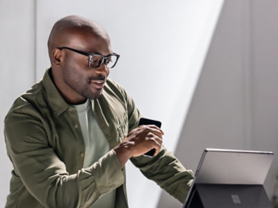A man in glasses, wearing a green jacket, smiles while using a laptop and smartphone.