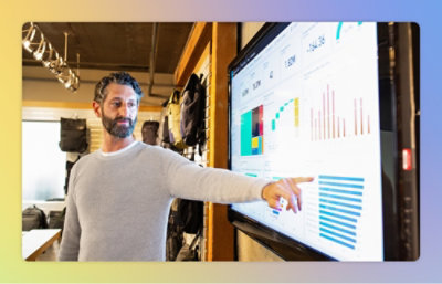 A man in a gray sweater points at statistics on a large screen displaying colorful graphs and charts.