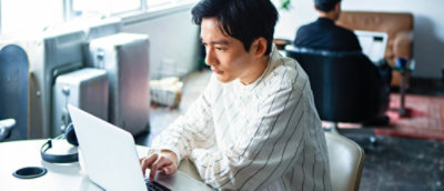 Person working on a laptop at a desk in a bright office, with another person working in the background.