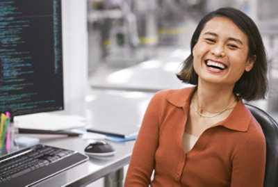 一個人微笑地坐在桌前，桌上有一部電腦，螢幕上顯示著數行的程式碼。