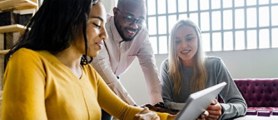 Trzy osoby zgromadzone wokół tabletu dyskutują o czymś, uśmiechając się. Siedzą w jasnym pokoju z dużymi oknami w tle.