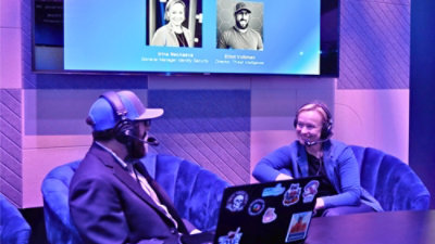 Studio panel discussion with two participants wearing headsets seated on chairs, using a laptop, with a presentation screen displayed behind them.