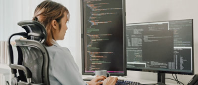 Woman in an office chair reviews code on dual computer screens in a bright workspace.