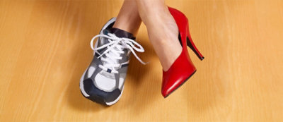 Jedna stopa w szarym sportowym trampku, a druga w czerwonym bucie na wysokim obcasie, stojące na drewnianej podłodze.