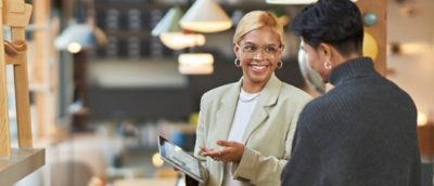 Two people standing indoors, engaged in conversation; one is holding a tablet. The background shows hanging lights and a modern workspace setting.